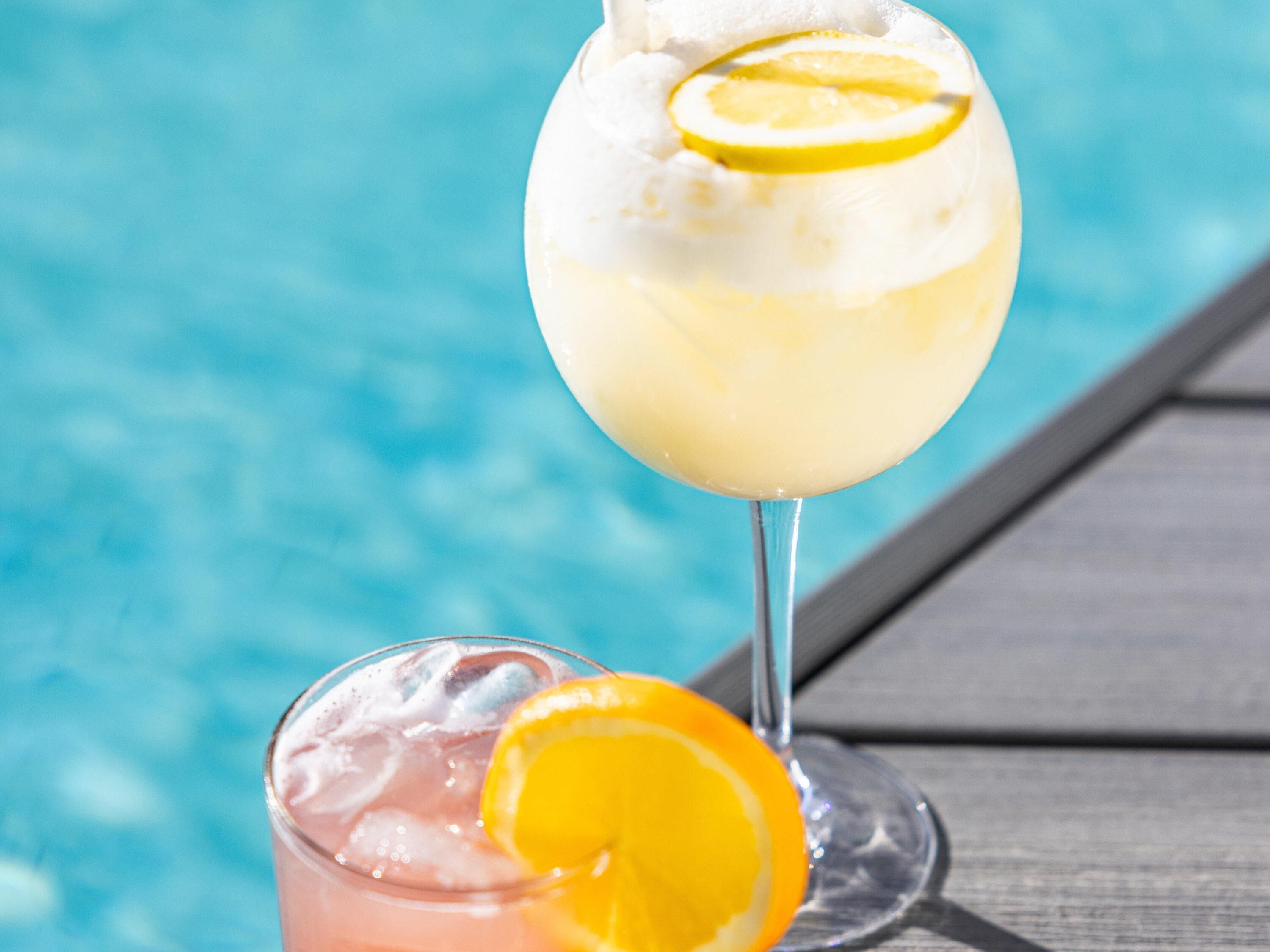 A poolside setting with a glass of a light-colored beverage garnished with a slice of lemon, and another glass with a slice of orange.