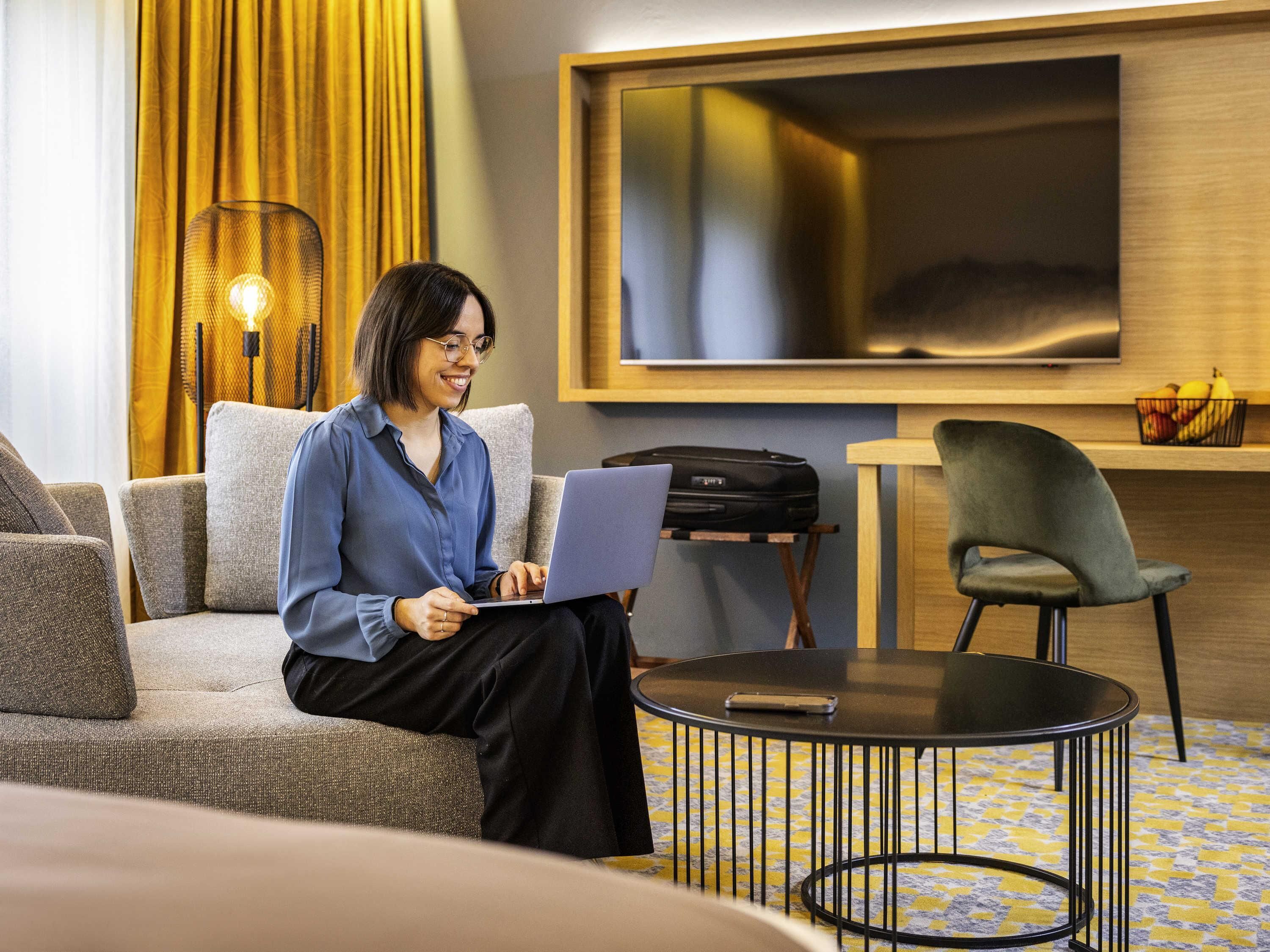 A person sitting on a sofa with a laptop, a suitcase on a small table, and a TV mounted on the wall.