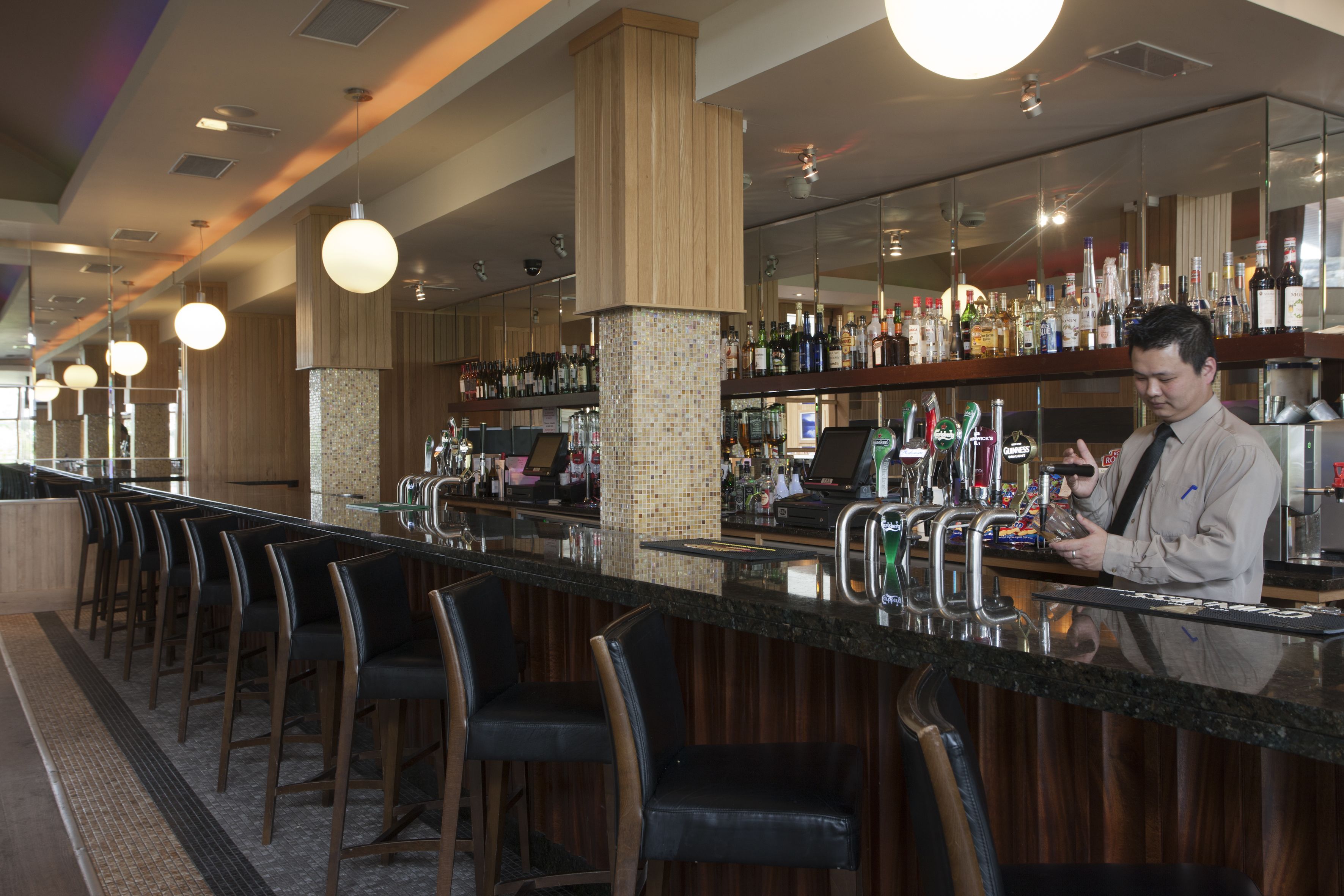 A bar area with a long counter, multiple bar stools, and a variety of bottles and glasses on shelves.