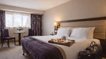 A hotel room with a large bed, a bedside table with a lamp, a chair, a small table with flowers, and a tray of pastries.