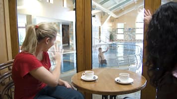 Una mujer sentada a una mesa con una taza de café, mirando un área de piscina donde un niño juega.
