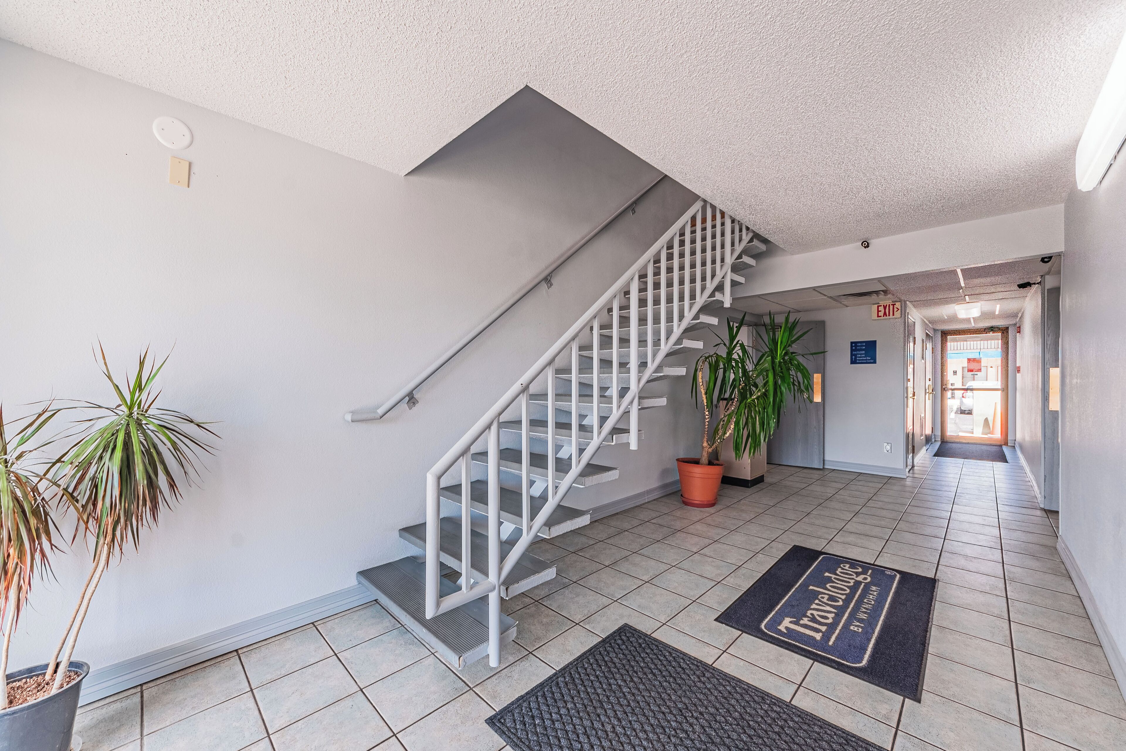 A modern hotel lobby with a staircase, potted plants, and a doormat with 'Travelodge' branding.