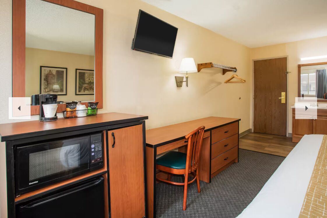 A hotel room with a desk, a chair, a bed, a television, a microwave, and a small kitchenette.