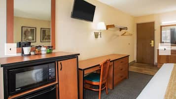 A hotel room with a desk, a chair, a bed, a television, a microwave, and a small kitchenette.