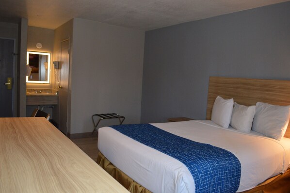 A hotel room with a bed, a wooden desk, a mirror, and a wall-mounted light fixture.