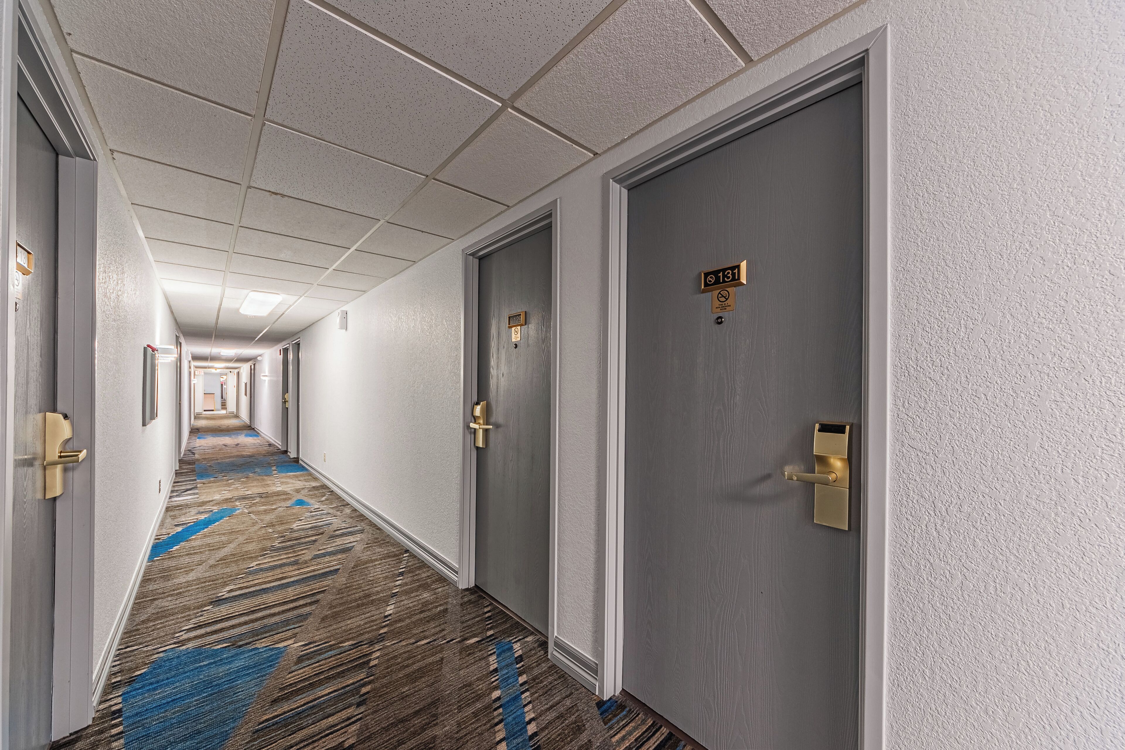 A hotel corridor with carpeted floors, doors with room numbers, and a ceiling with recessed lighting.