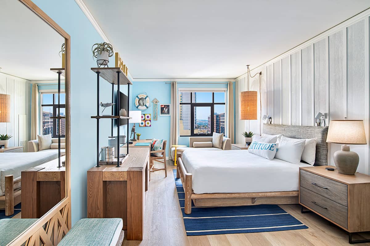 A modern hotel room with a large bed, a desk, a chair, and a bookshelf.