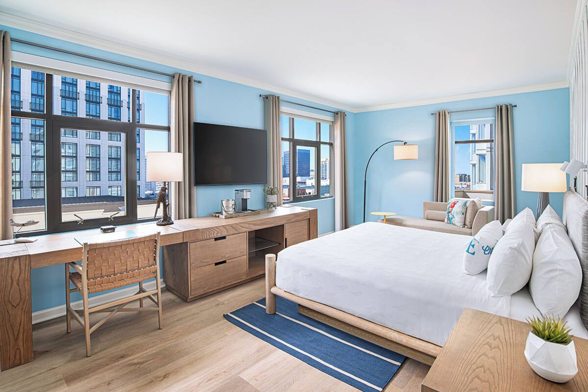A hotel room with a large bed, a desk with a computer, a TV, and a view of the city through large windows.