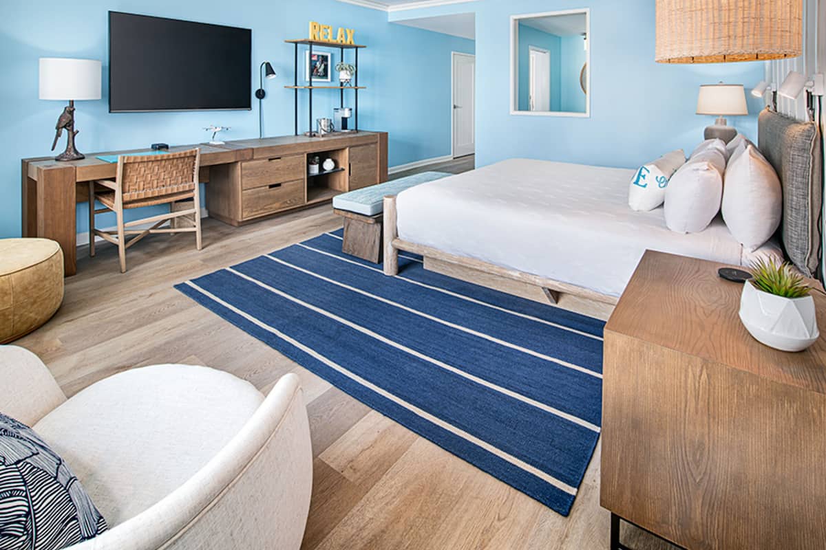 A modern hotel room with a large bed, a desk, a TV, and a blue-striped rug.