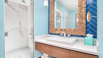 A bathroom with a walk-in shower, a large mirror with a woven frame, a white sink, and a wooden vanity.