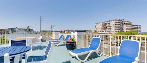 Eine Dachterrasse mit Liegestühlen und Tischen, einem Blumentopf und Blick auf ein mehrgeschossiges Gebäude.
