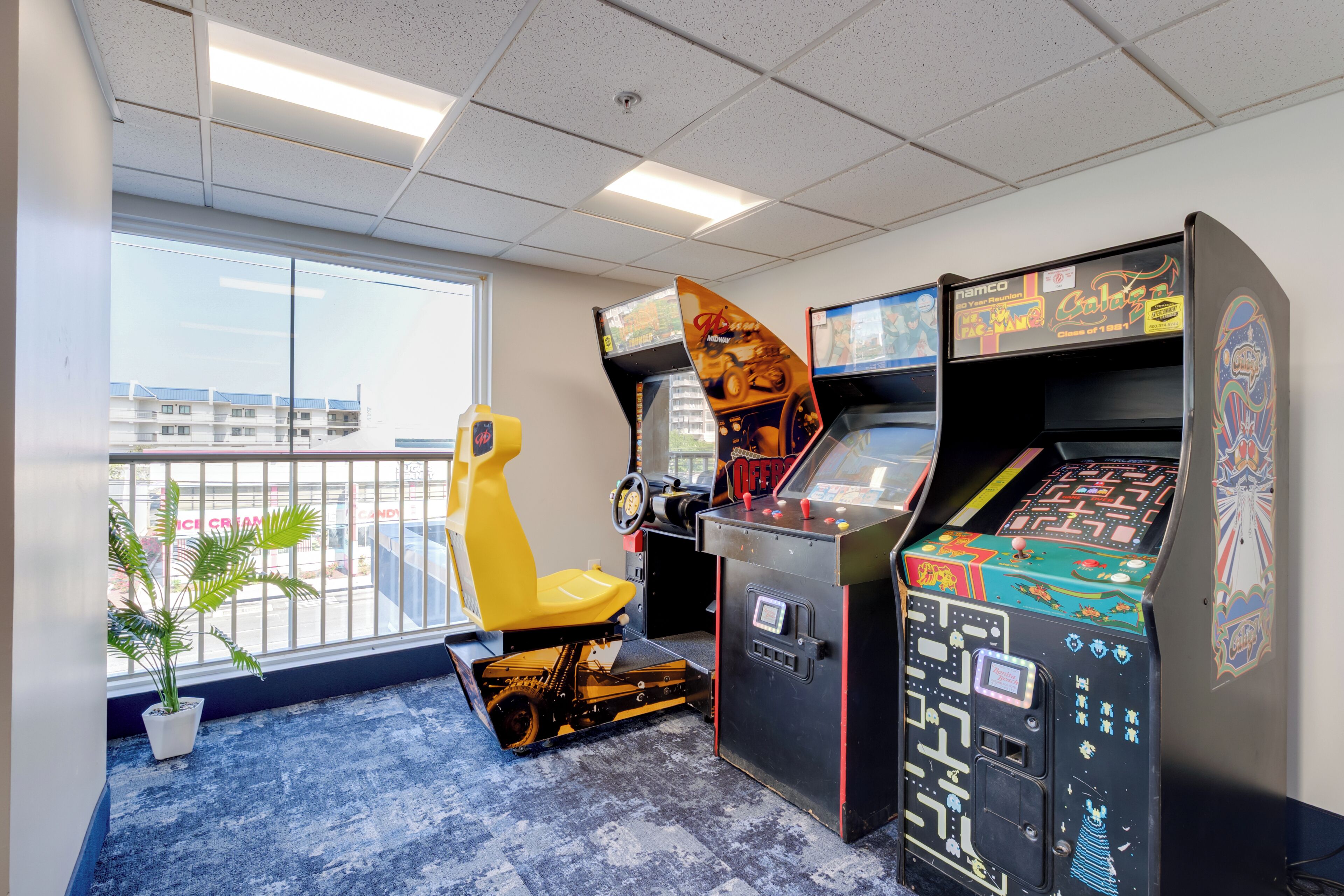 A room with arcade games, a yellow chair, a potted plant, and a view of buildings outside.