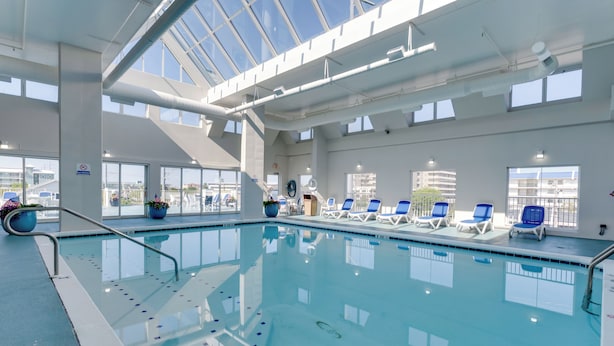 An indoor swimming pool with clear blue water, sunlit by skylights, surrounded by lounge chairs and potted plants.