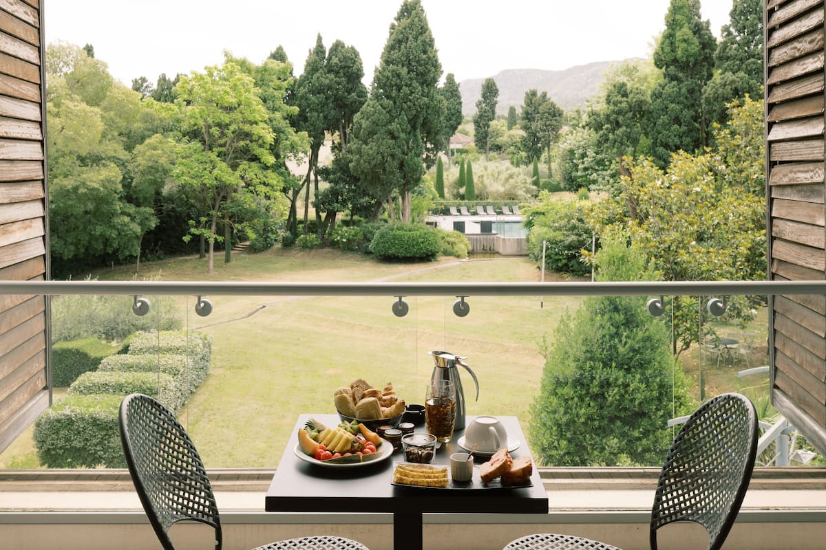 A balcony with a table set for breakfast, overlooking a well-manicured garden and a pool.