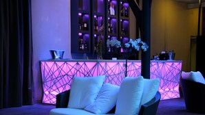 Una zona de bar moderna amb un taulell de bar il·luminat amb làmpades de neó, seients amb coixinets blancs i una taula de vidre.