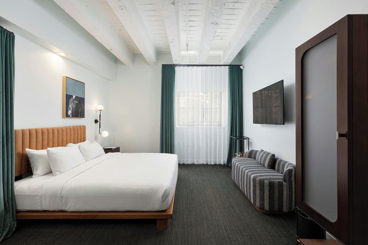 Una habitación de hotel con cama, sofá, televisor y ventana con cortinas.
