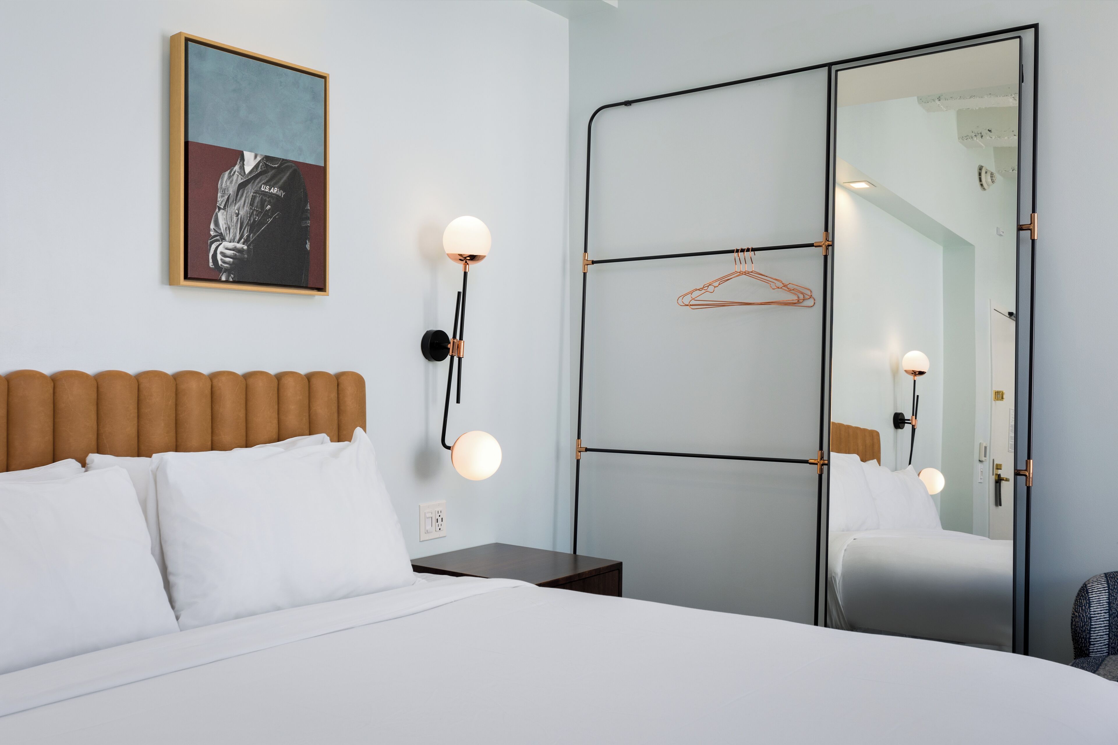Habitación de hotel con cama, mesita de noche, lámpara de pared, cuadro enmarcado y espejo con perchero.