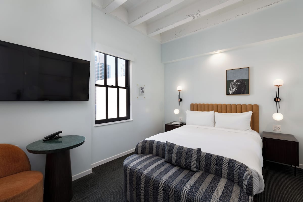 Habitación de hotel con cama, televisión montada en la pared, una mesita pequeña y un banco.
