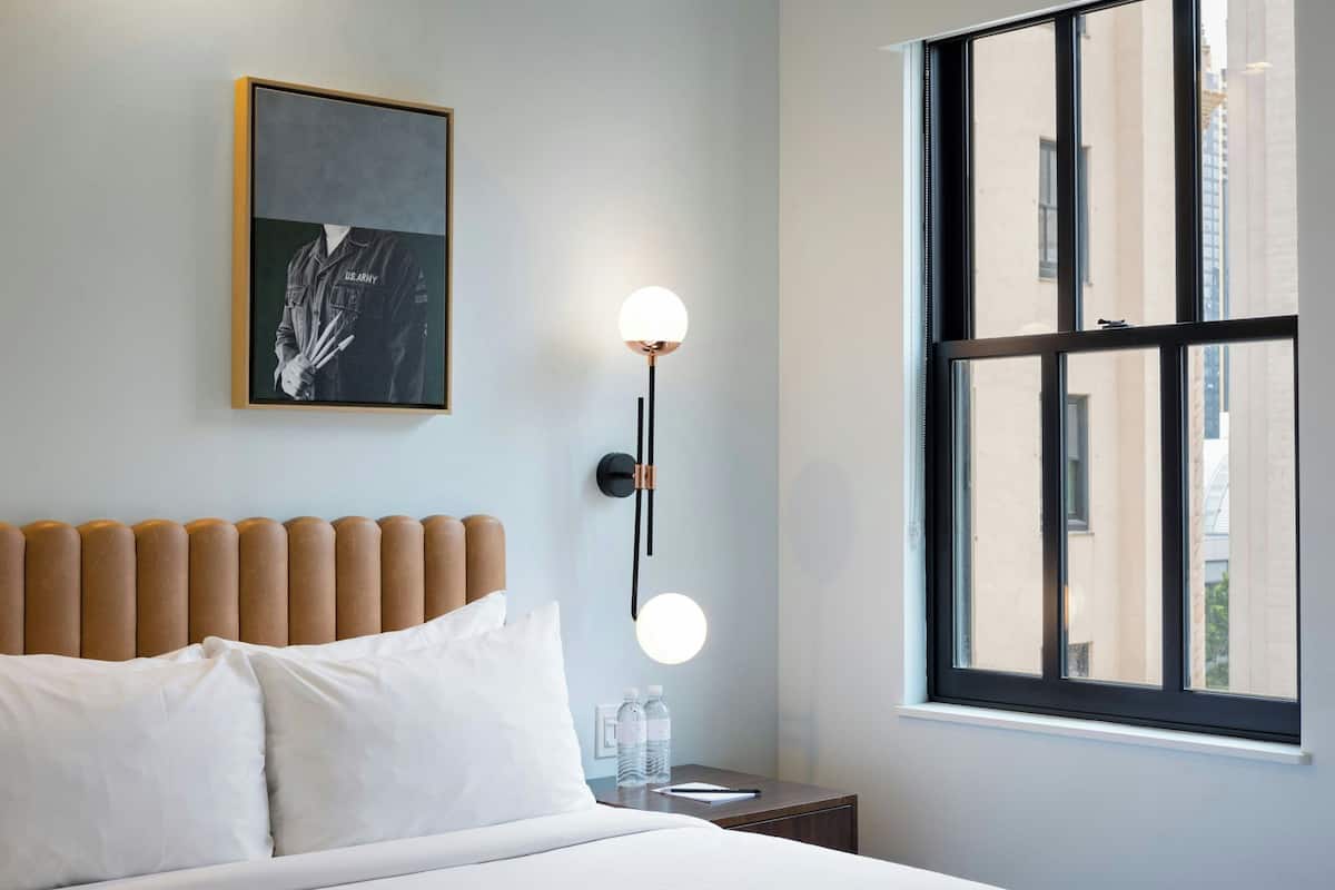 Habitación de hotel con una cama, una mesita de noche con lámpara, un cuadro enmarcado en la pared y una ventana con vistas a edificios.