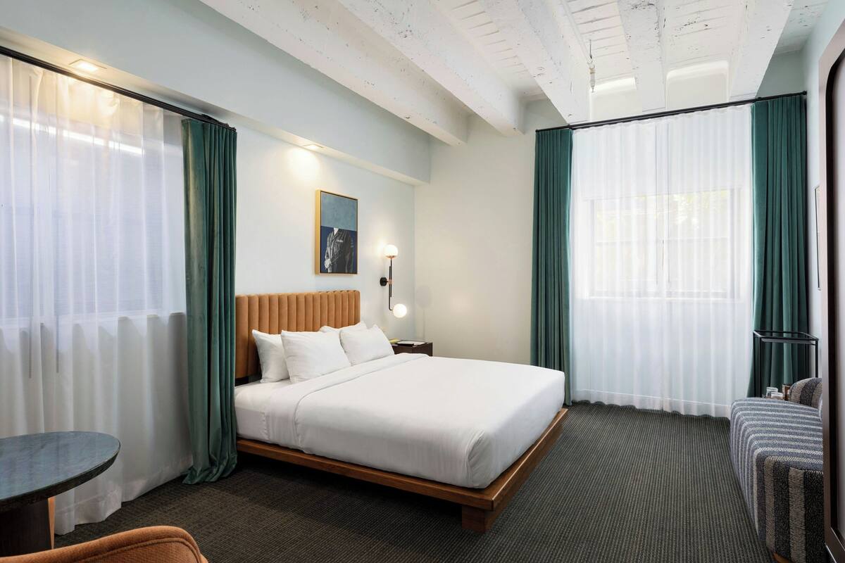 Habitación de hotel con una cama grande, un escritorio y una silla. Hay una ventana con cortinas y un cuadro en la pared.