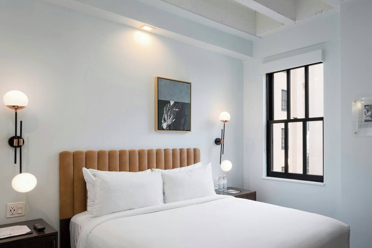 Una habitación de hotel moderna con una cama grande, cabecera, lámparas de noche, una ventana y una obra de arte enmarcada en la pared.
