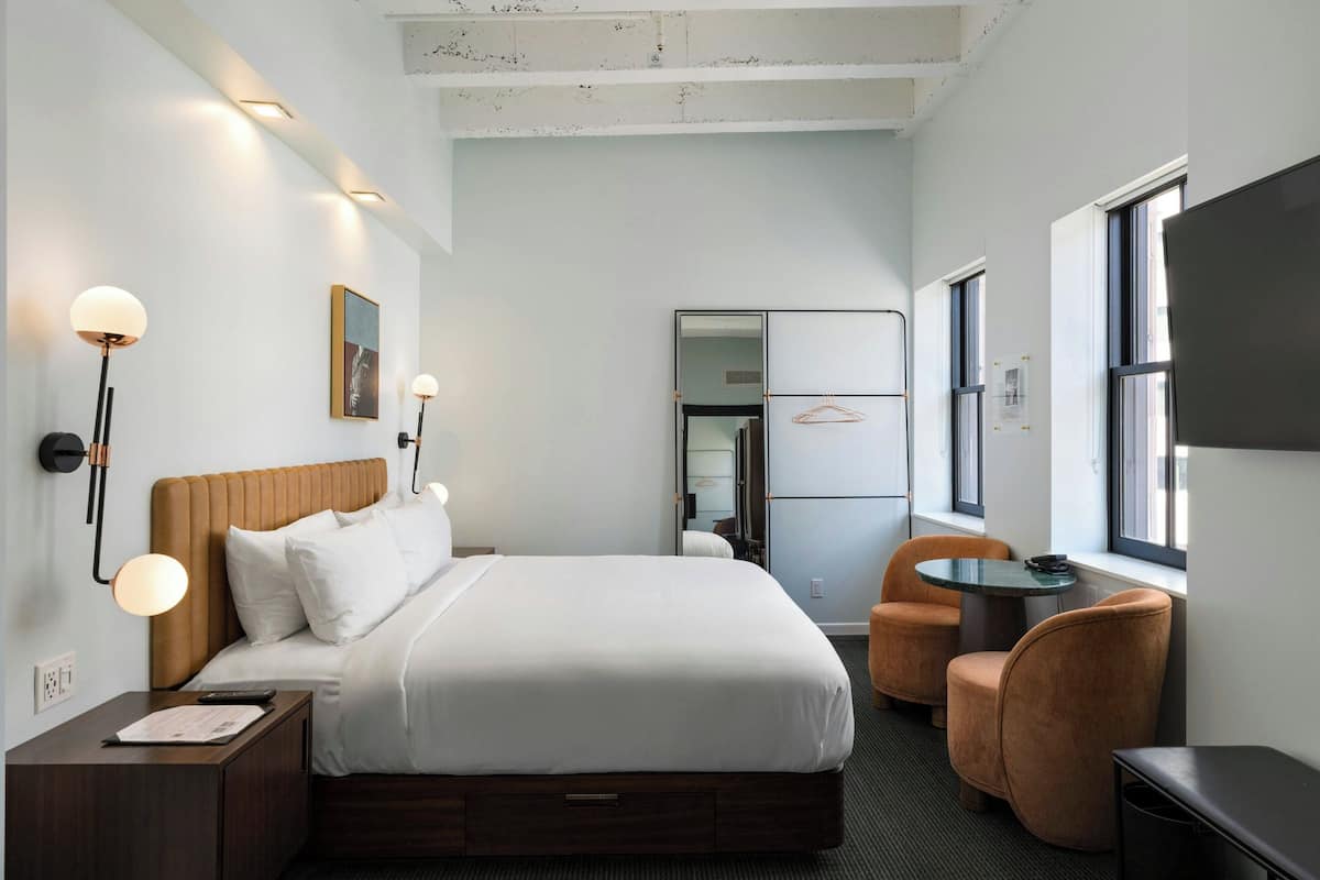 Una habitación de hotel con cama, mesita de noche, lámpara, silla y una pequeña mesa.