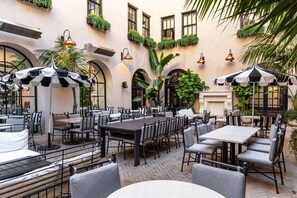 Café al aire libre con mesas y sillas, rodeado de plantas y elementos arquitectónicos.