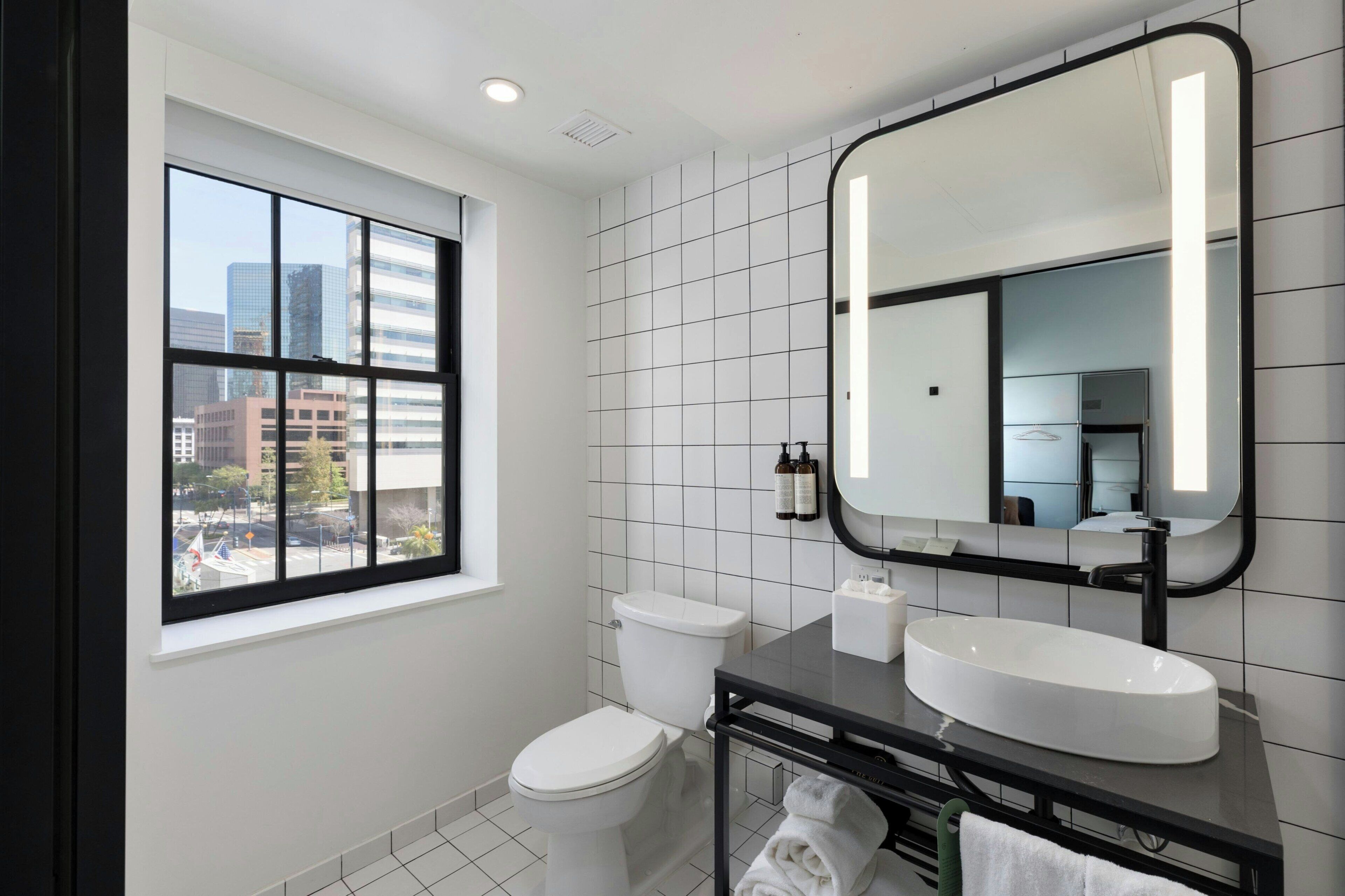Un baño moderno con un ventanal grande, un inodoro blanco, un tocador negro con lavamanos blanco y un espejo con marco negro.