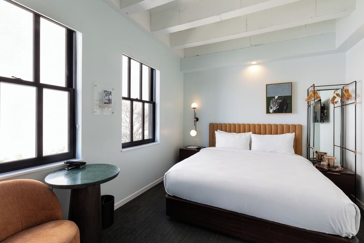Habitación de hotel con una cama grande, una mesa redonda, una silla y una ventana con persianas.