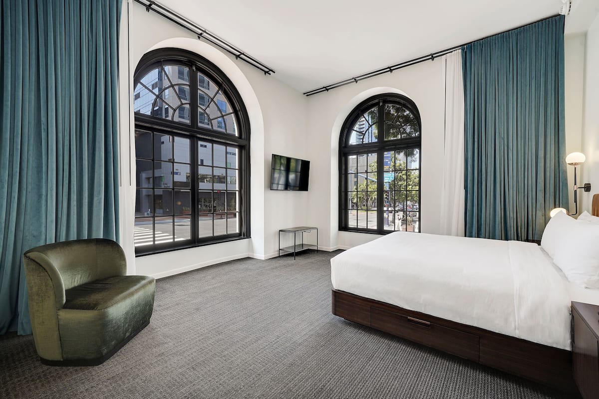 Habitación de hotel con una cama grande, una silla, un televisor y dos ventanas amplias con cortinas.