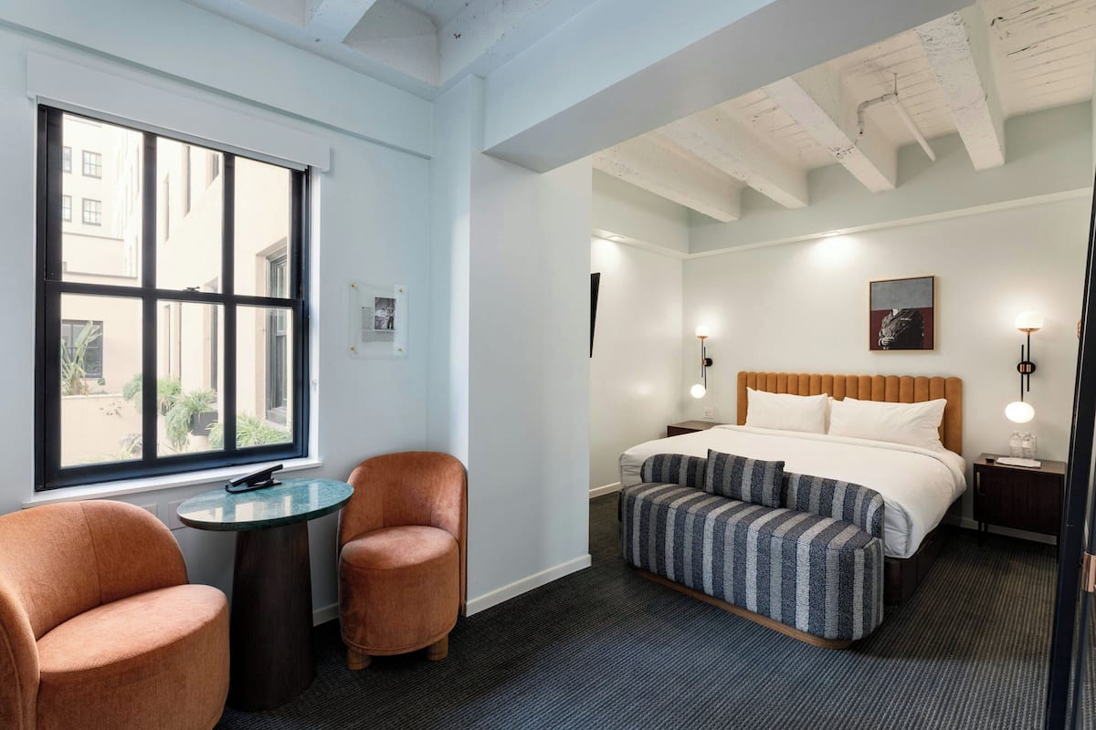 Habitación de hotel con un ventanal grande, una cama con ropa de cama a rayas, una mesita redonda pequeña, dos sillones y una mesita de noche con lámpara.