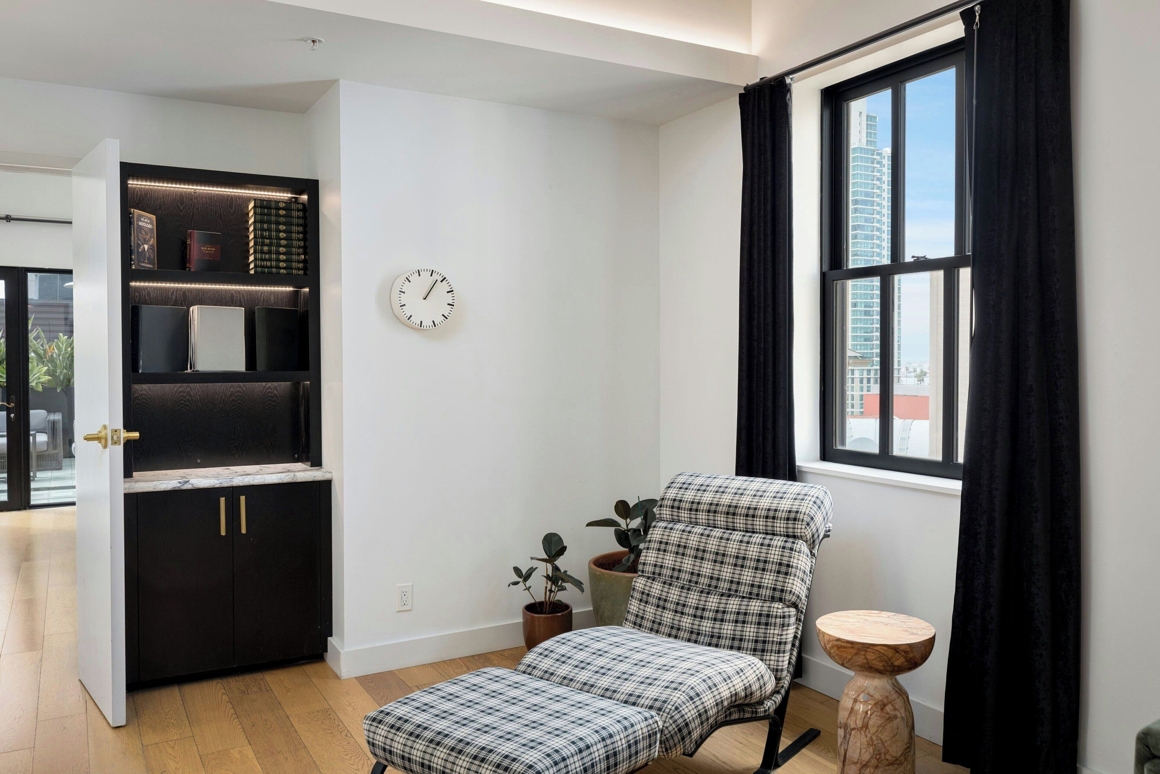 A modern living room with a black and white checkered armchair, a small round table, a bookshelf with books, a clock, and a window with curtains.