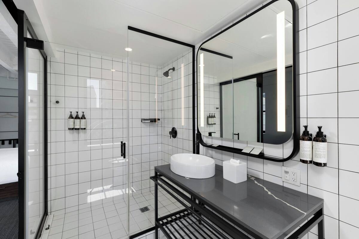 Un baño moderno con un espejo grande, azulejos blancos, una encimera negra y una ducha con paredes de vidrio.
