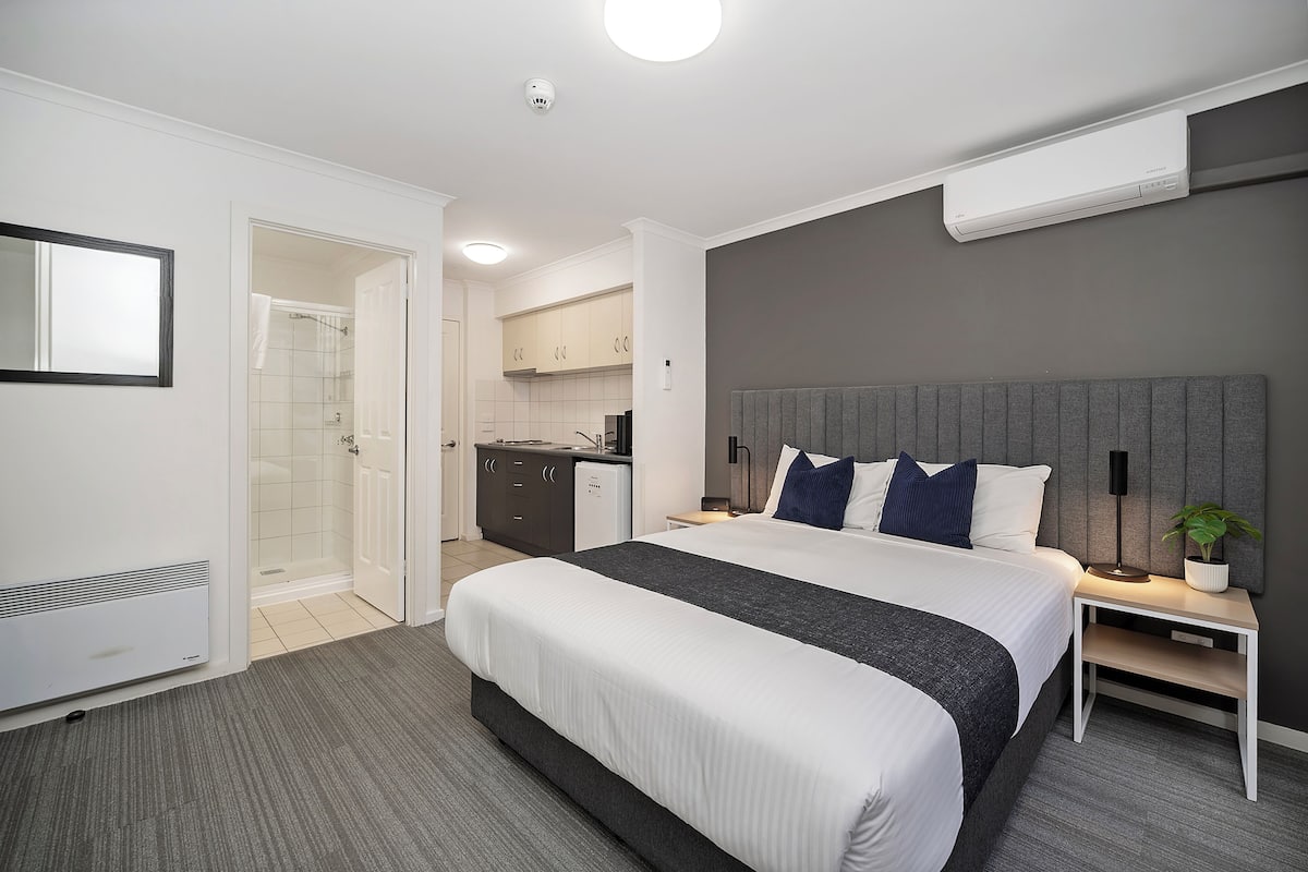 Una habitación de hotel moderna con una cama grande, una mesita de noche con lámpara, un escritorio integrado, zona de ducha y una kitchenette.