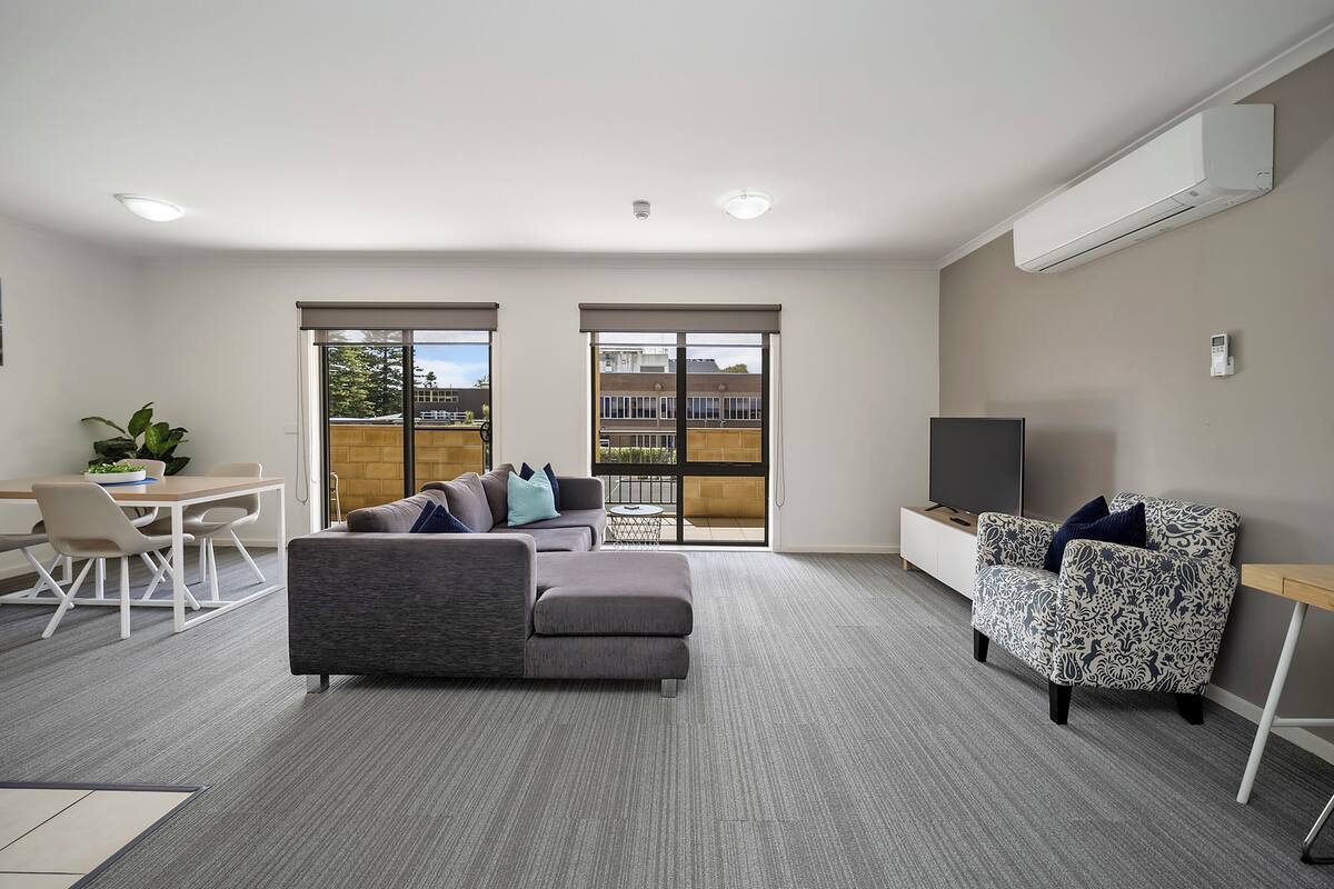 Una sala moderna con un sofá seccional gris, un sillón estampado, un televisor y un comedor con mesa y sillas.