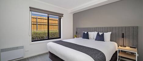 A bedroom with a large bed, a bedside table, a window with a view of a fence, and a ceiling light.