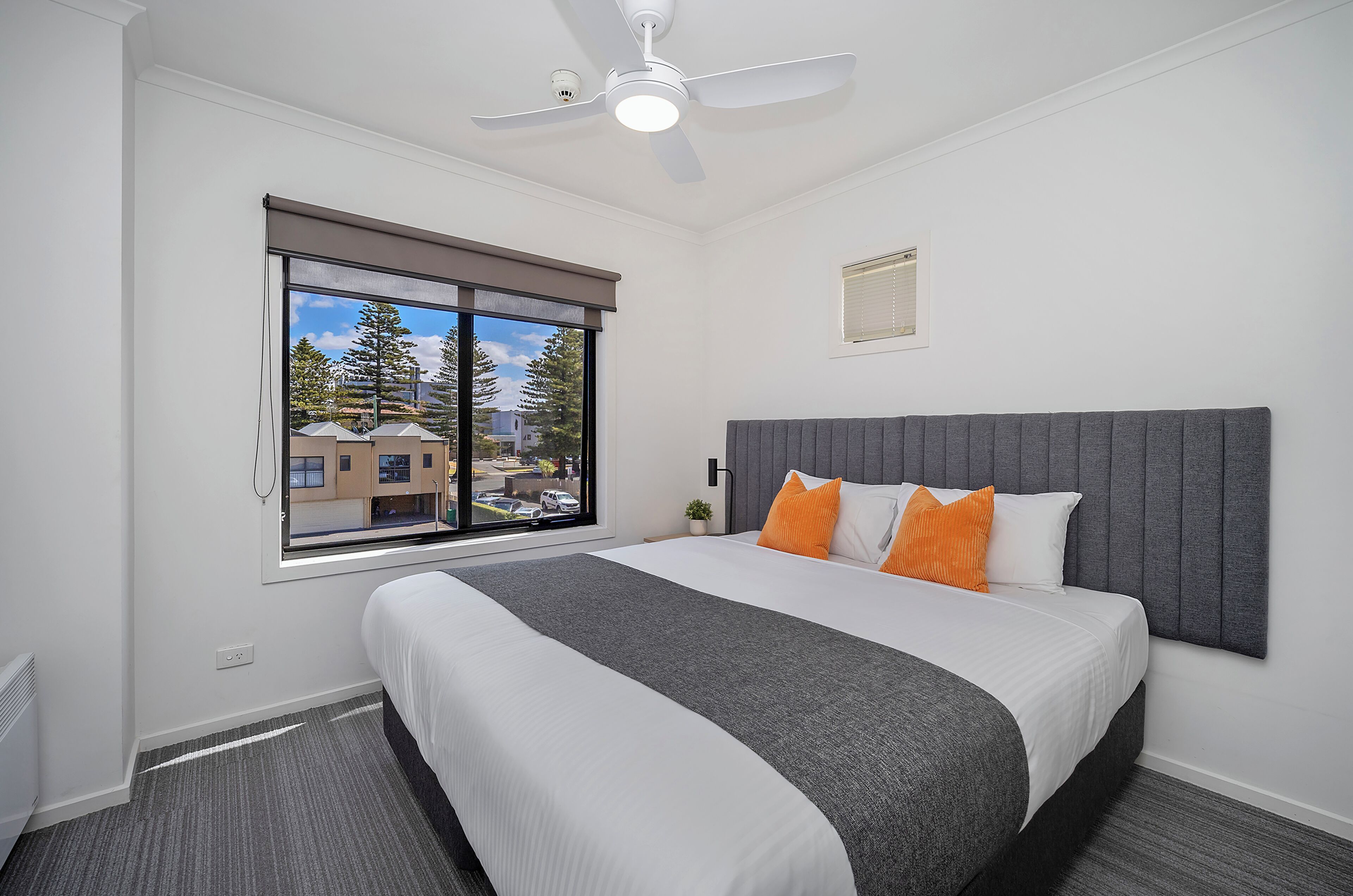 A hotel room with a bed, a ceiling fan, a window with a view, and a small plant.