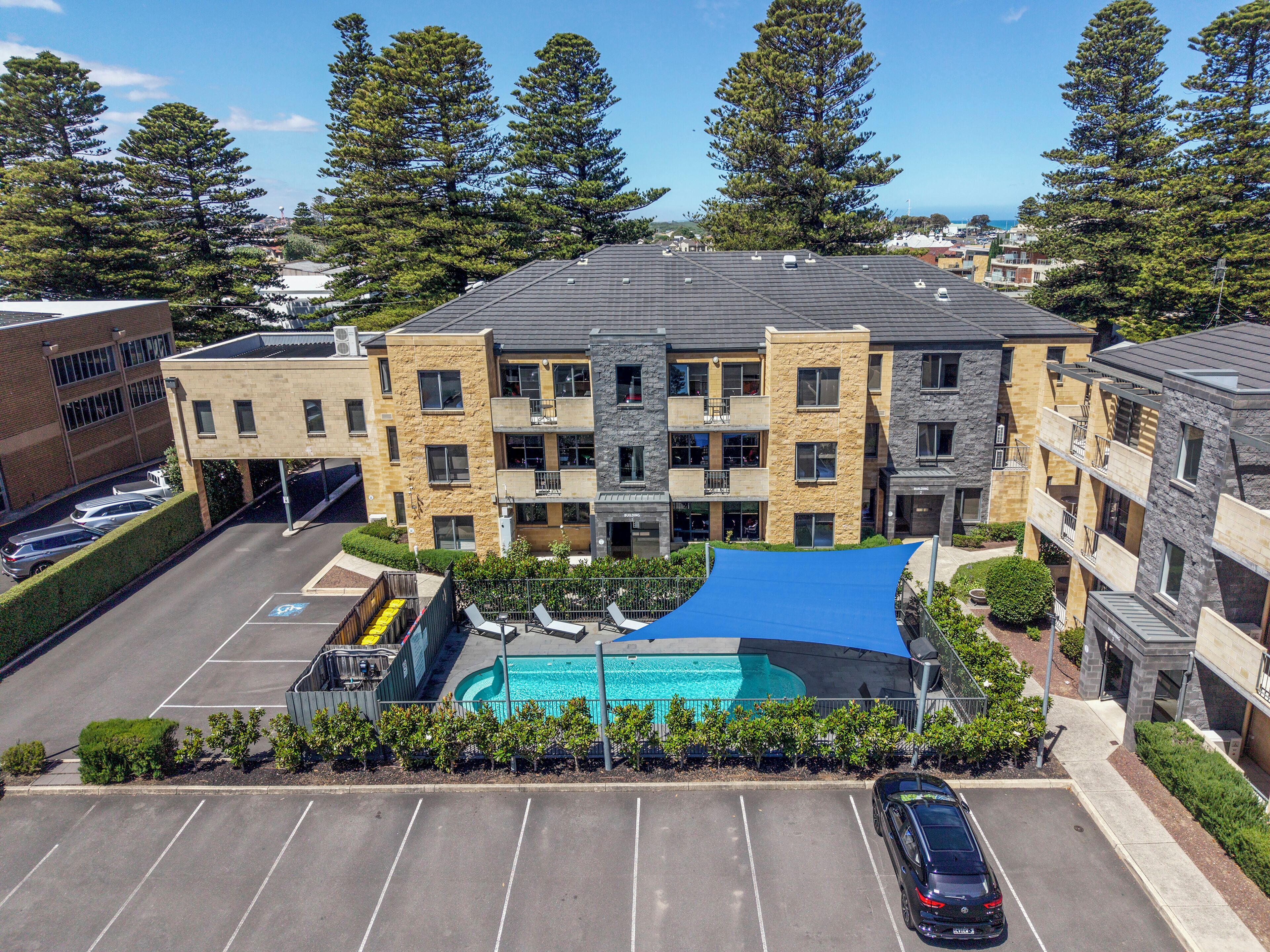A multi-story building with a swimming pool, parking lot, and surrounding greenery.