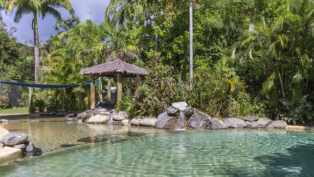 Piscina límpida com pedras e uma construção de telhado de palha, tudo envolto em vegetação exuberante.