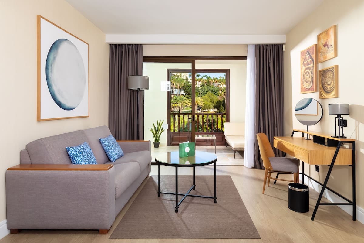 Ein modernes Wohnzimmer mit Sofa, Glastisch, Schreibtisch und Stuhl sowie Blick auf einen Balkon mit Pflanzen.