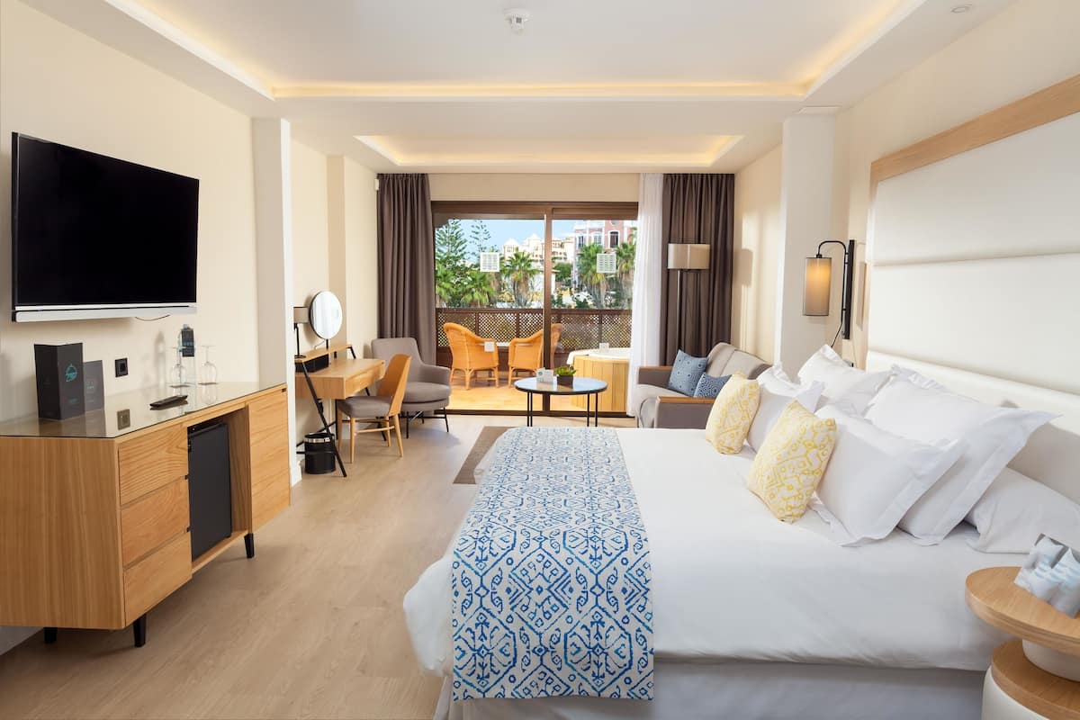 Ein Hotelzimmer mit einem großen Bett, einem Schreibtisch mit Fernseher, einer Couch und einem Balkon mit Sitzgelegenheit.