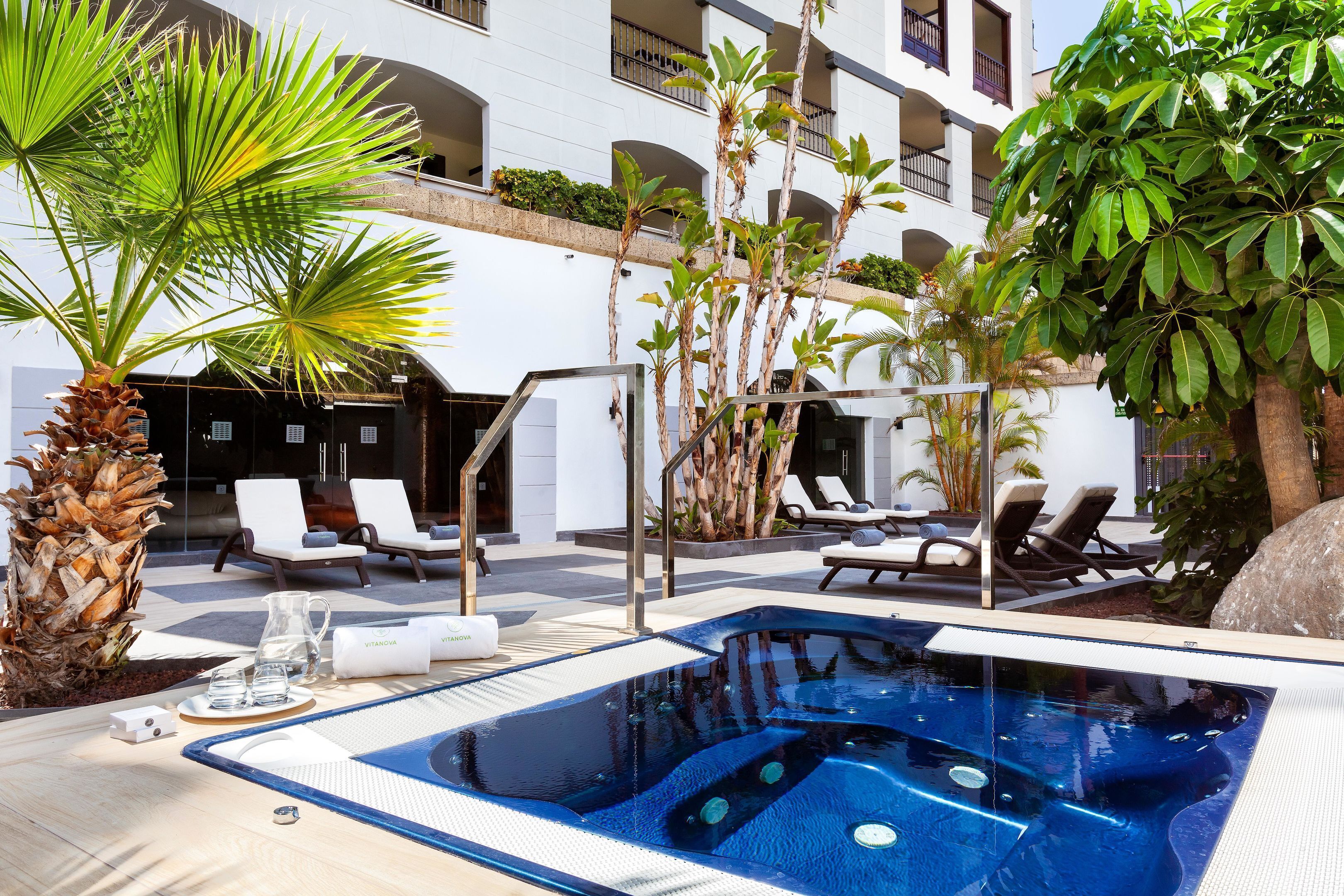 Piscina con sillones de descanso y palmeras en un patio de hotel.