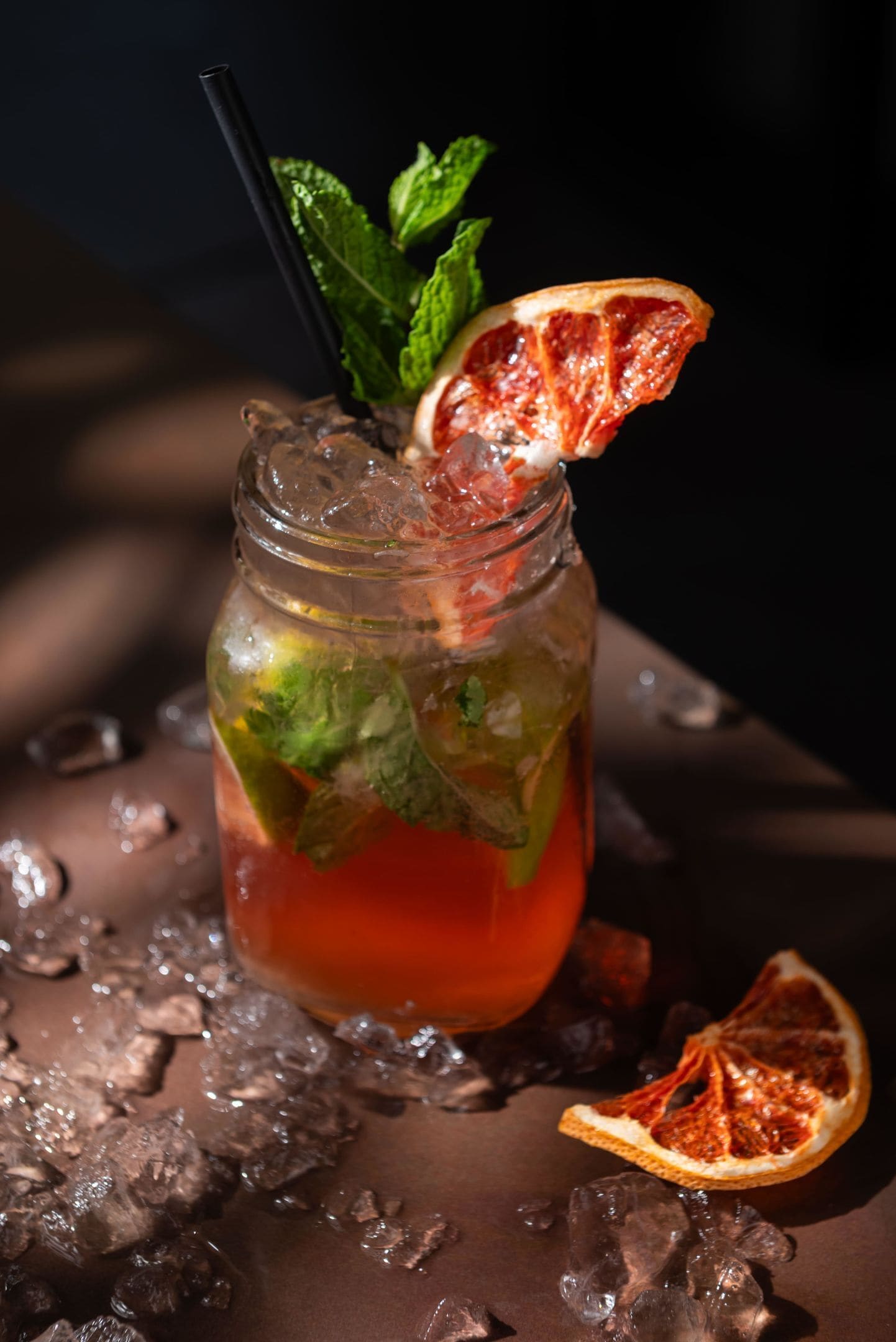 Un verre avec une paille noire, décoré de feuilles de menthe et d’une tranche d’orange sanguine, entouré de glaçons.