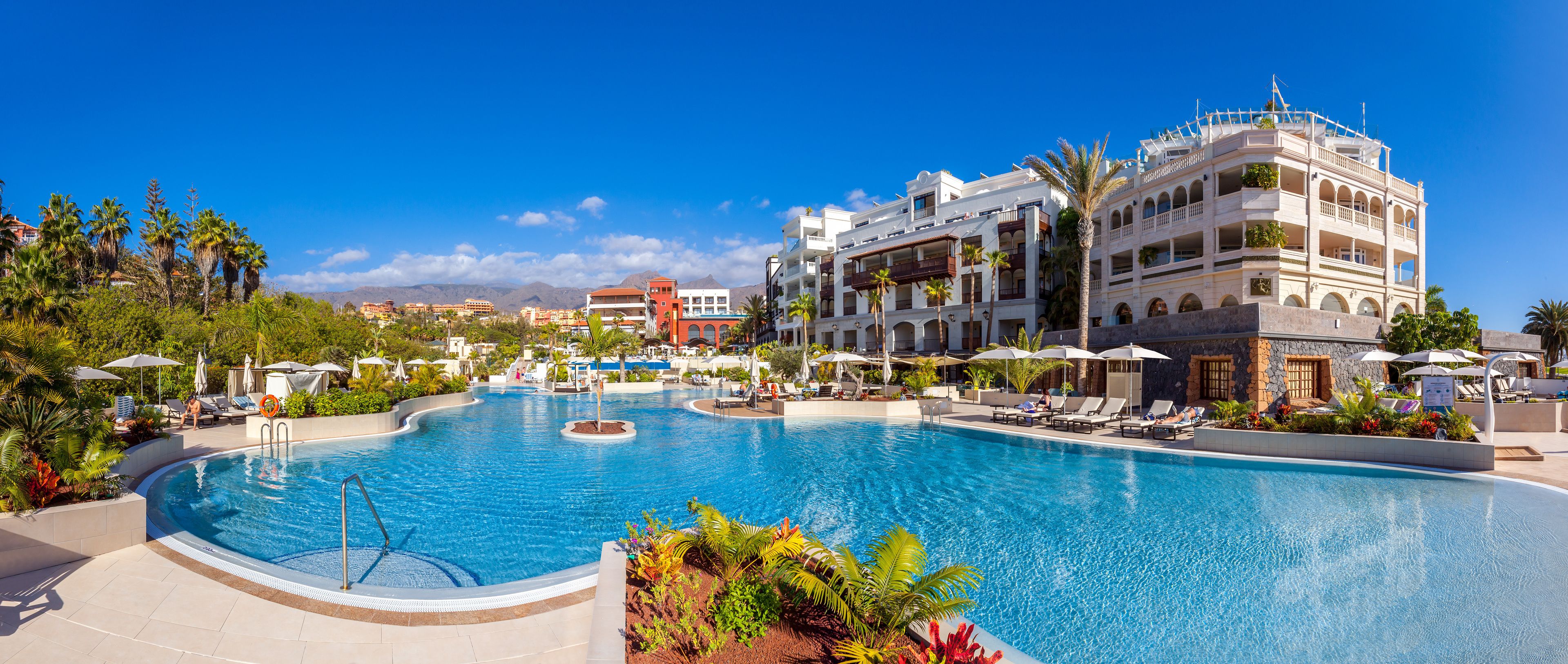 A large swimming pool surrounded by lounge chairs and umbrellas, with a hotel building in the background.