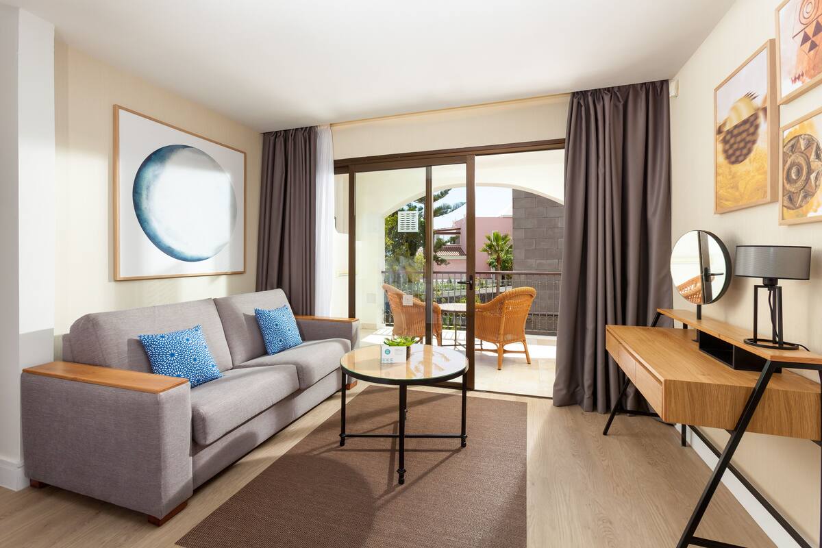 Ein modernes Hotelzimmer mit grauem Sofa, einem Holztisch, einem Glastisch und einem Balkon mit Korbstühlen.