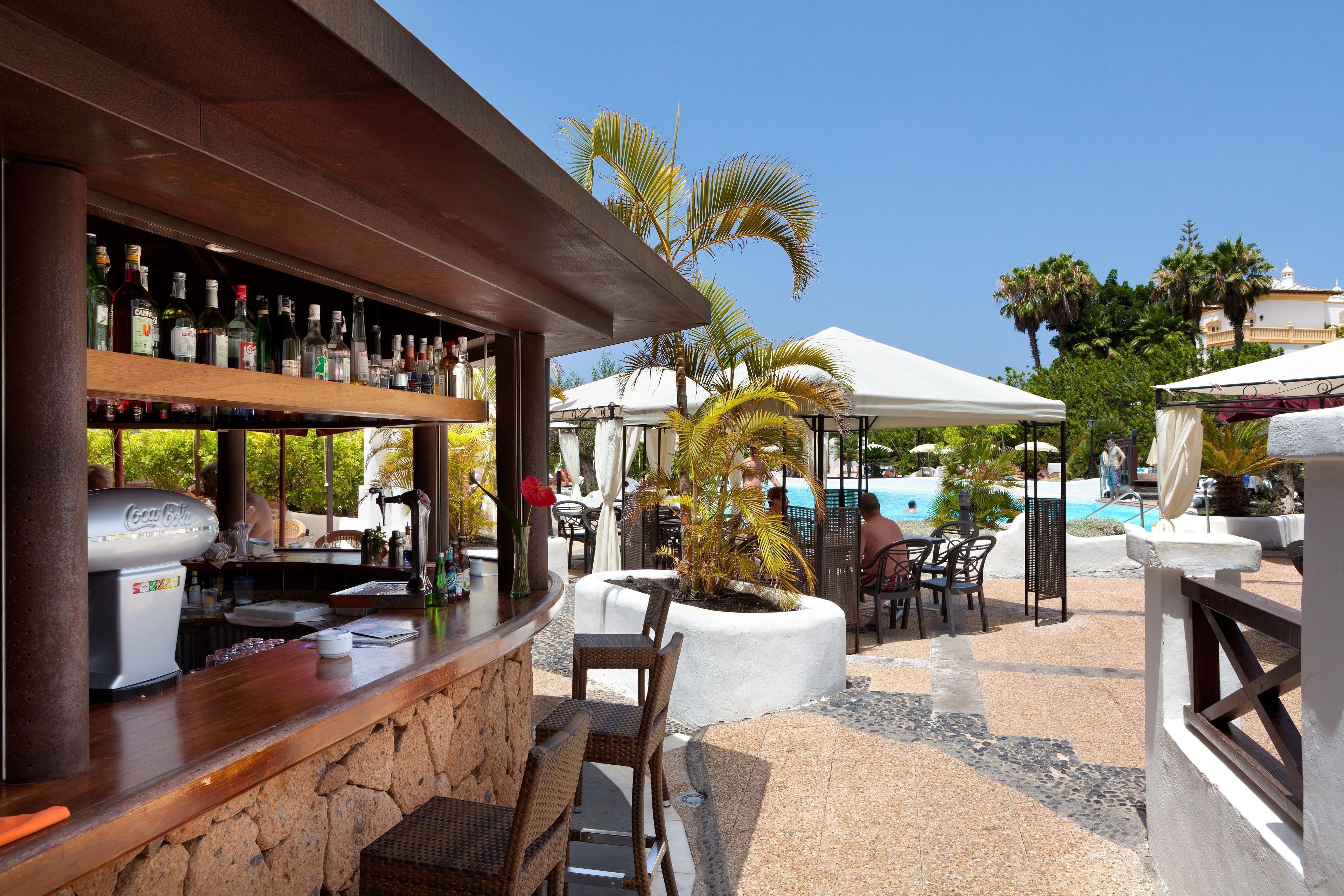 Uma área de bar com balcão de pedra, geladeira Coca-Cola e diversos frascos. Há áreas de estar ao ar livre com cadeiras de vime e guarda-sóis, e uma área de piscina ao fundo.