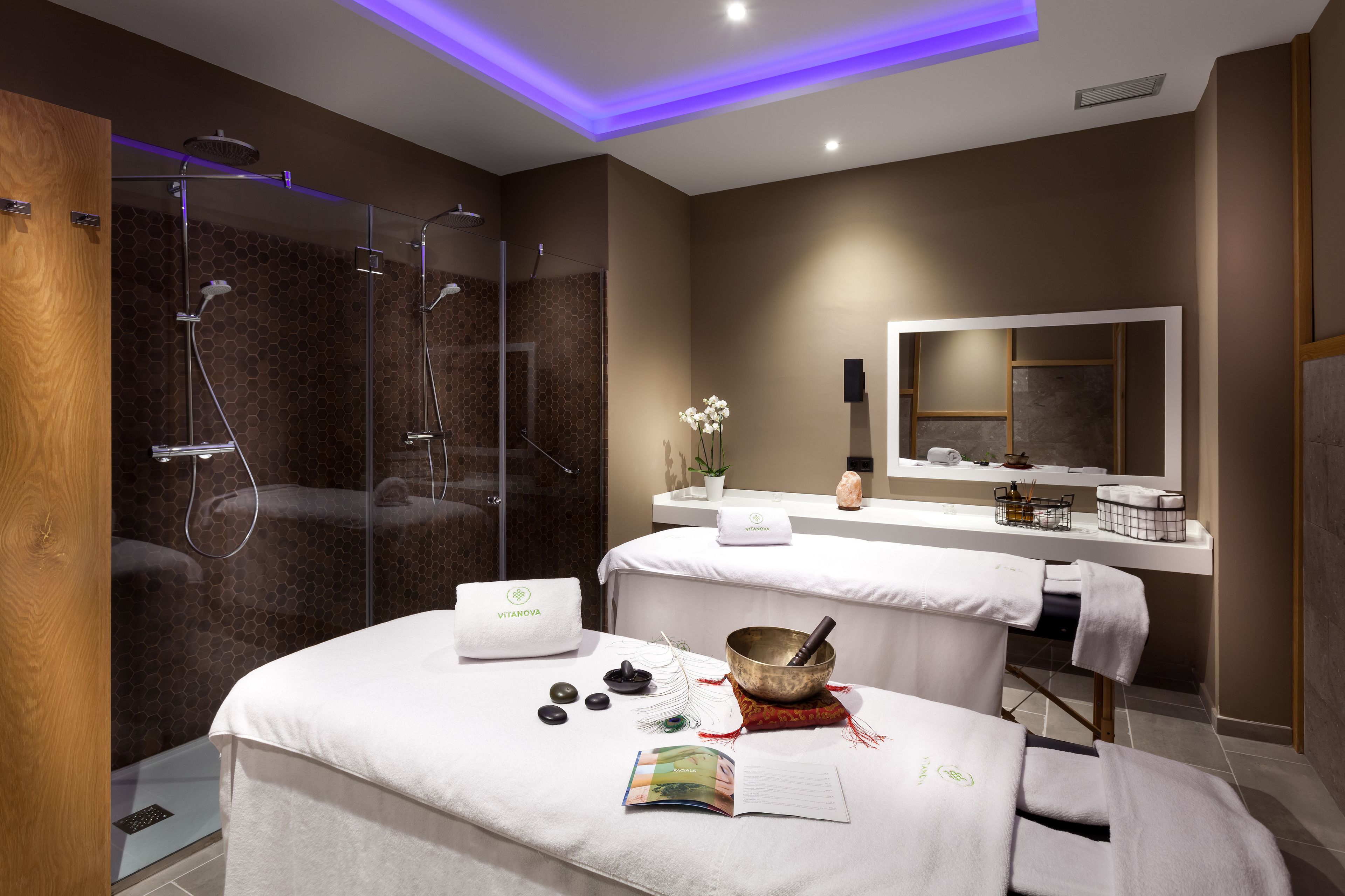 Un spa moderno con ducha de vidrio, camilla de masaje, zona de meditación con piedras y tocador con espejo y artículos de aseo.