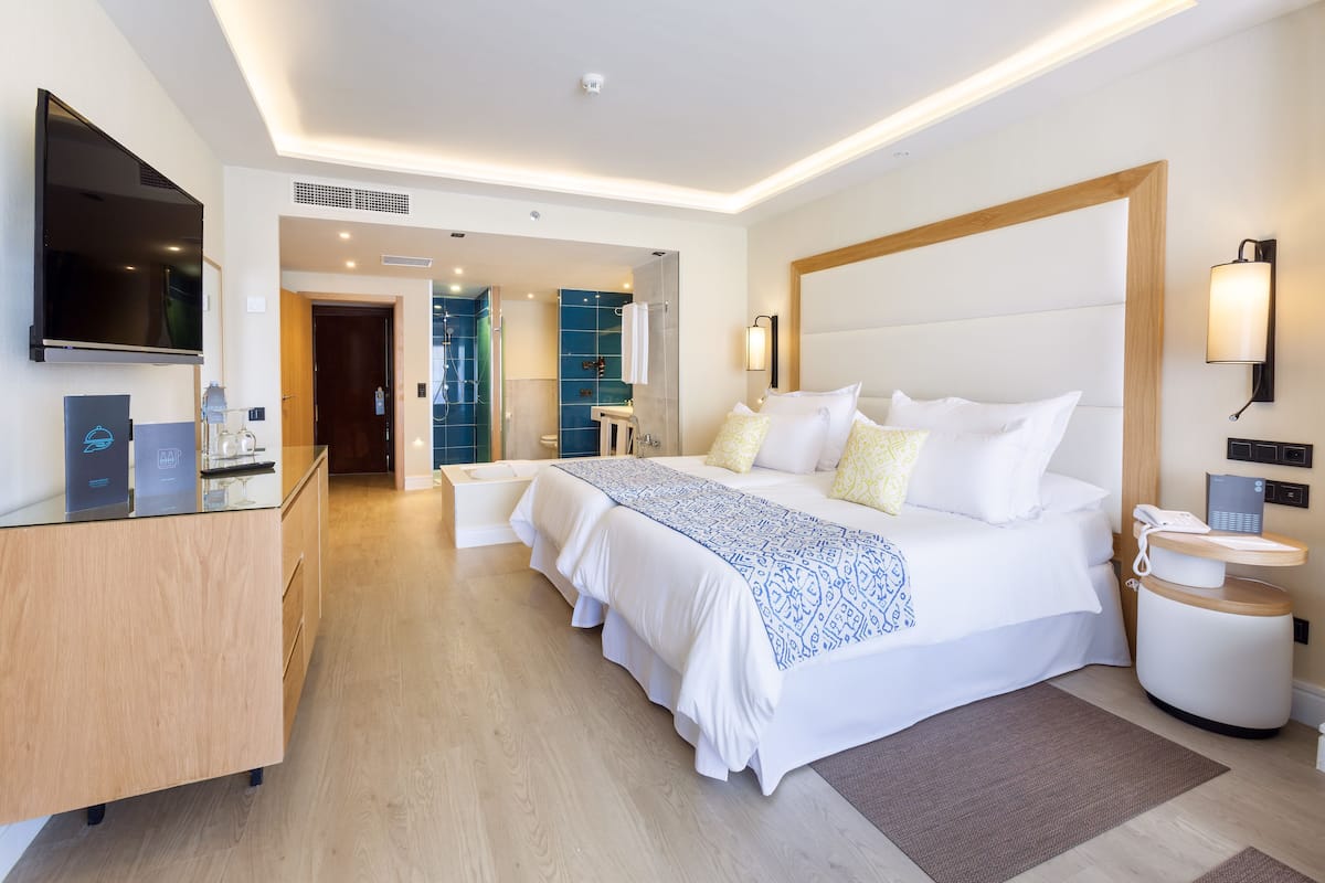 Ein modernes Hotelzimmer mit einem großen Bett, Nachttischen, einem Fernseher und einem durch eine offene Tür einsehbaren Badezimmer.