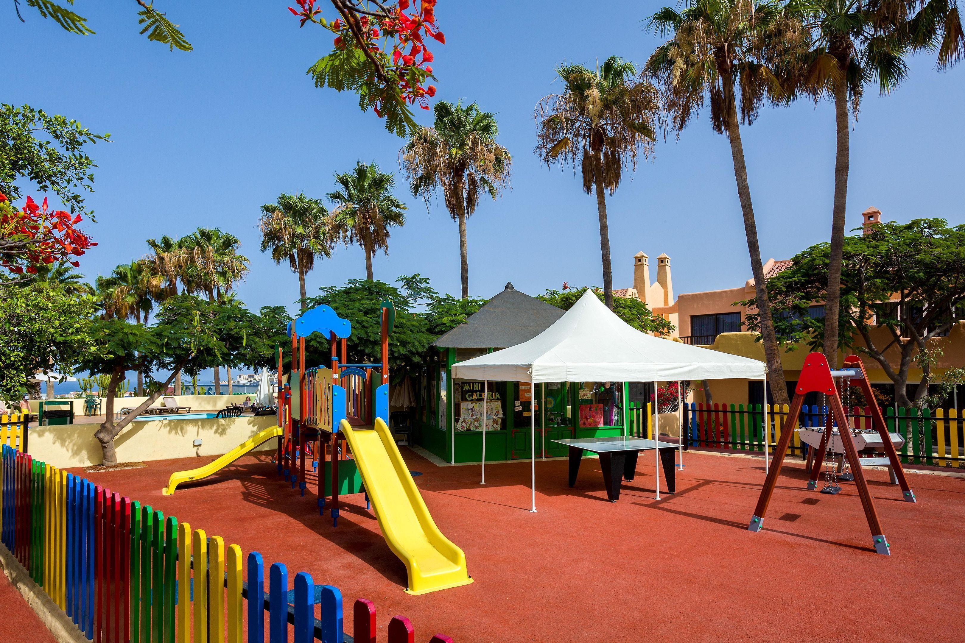 A playground with a yellow slide, a red swing set, and a white tent.
