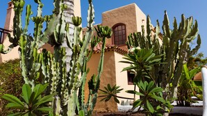 Un bâtiment à la façade beige et au toit recouvert de tuiles rouges, entouré de différentes espèces de cactus et de plantes succulentes.